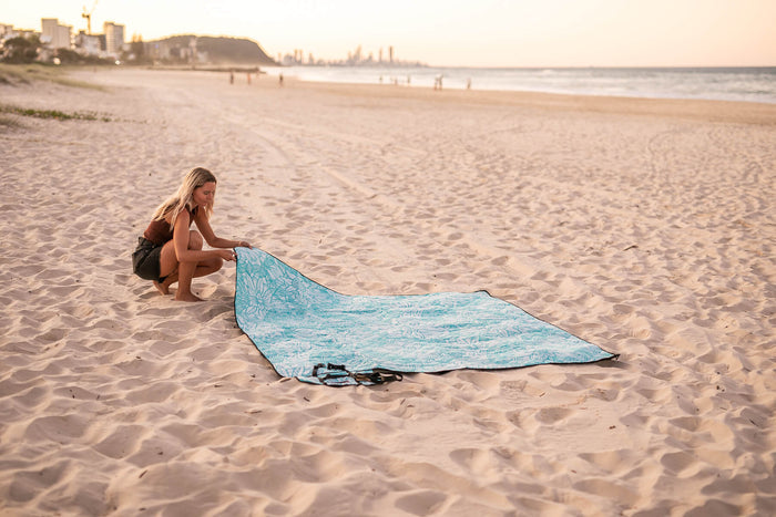 Turquoise patterned recycled picnic blanket laid on sand with woman crouching beside it at beach