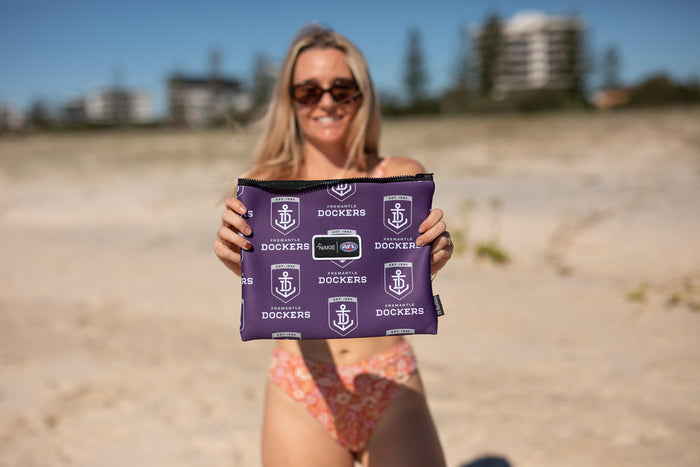 Purple Fremantle Dockers AFL beach towel with repeated anchor logo and text pattern, displayed at the beach.