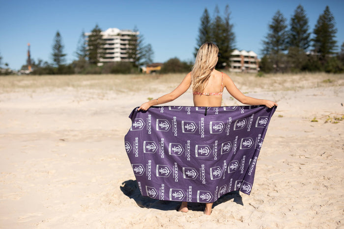 Purple Fremantle Dockers beach towel with repeating logo print, held open on sandy beach