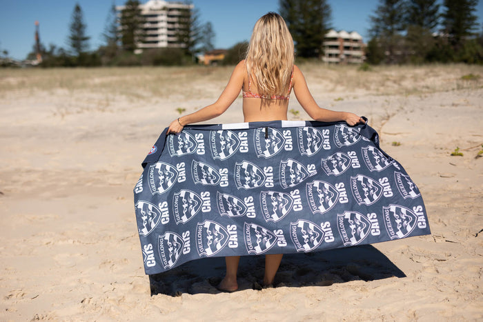 Geelong Cats navy beach towel with repeated white logo pattern, held by person on sandy beach