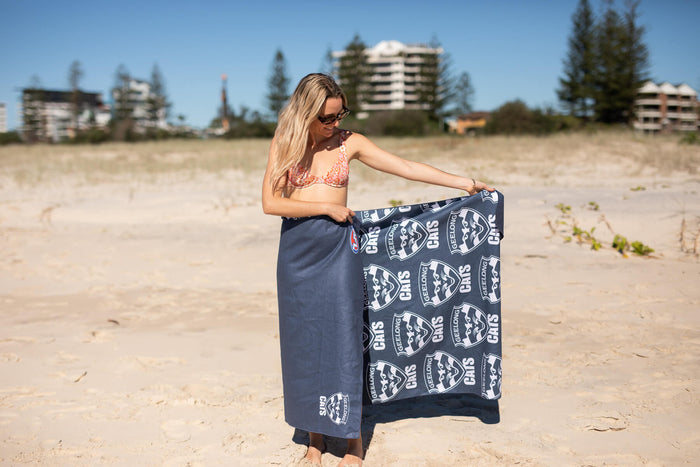 Navy Geelong Cats AFL beach towel with white logo repeat pattern, displayed on sand at beach.