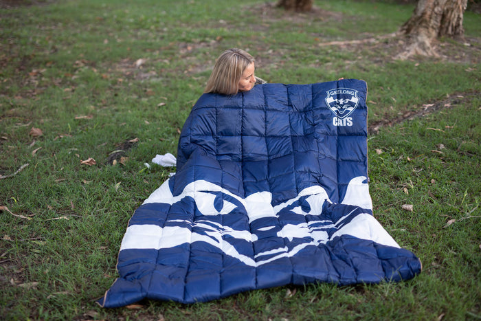 Navy Geelong Cats puffy blanket with white stripes, laid out on grass with woman wrapped in it