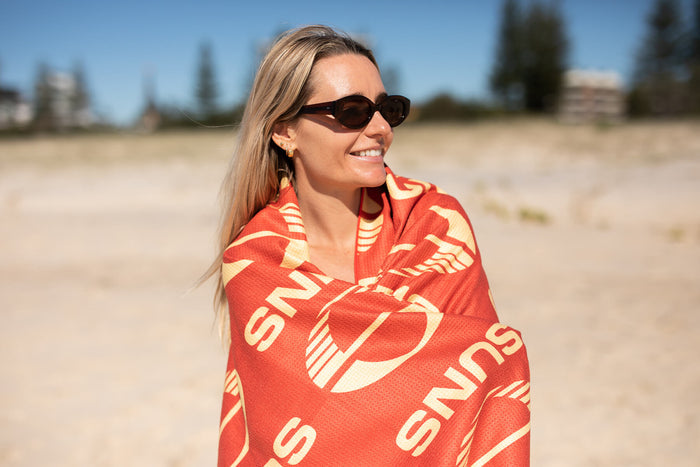 Red and gold Gold Coast SUNS AFL recycled beach towel wrapped around woman at beach, sand-free weave visible