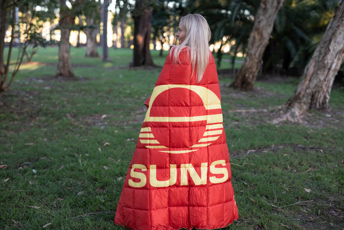 Red and gold quilted puffy blanket with Gold Coast Suns AFL logo and sun design, worn as a wrap outdoors.