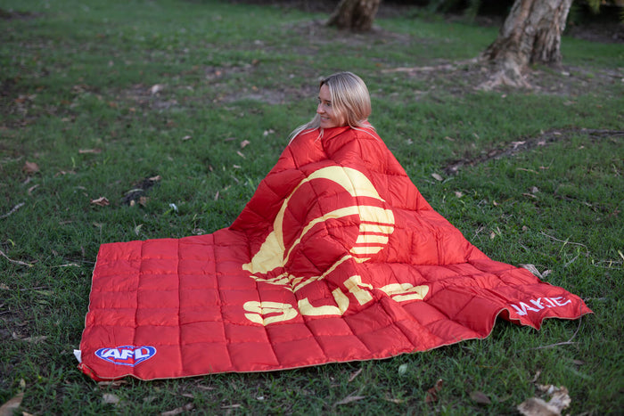 Red quilted puffy blanket with gold sun logo and AFL branding, displayed on grass with person wrapped inside.