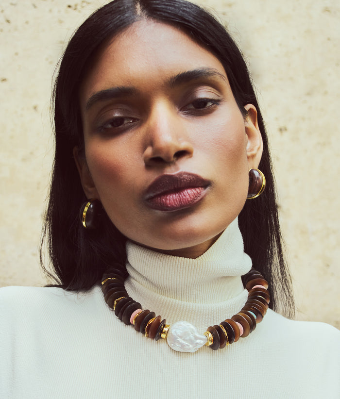 Model on tan stone backdrop wears white knit top with necklace and Halo Hoops in Acacia Wood