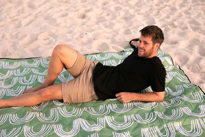 Green and white geometric picnic blanket with recycled material, displayed on sand with person reclining