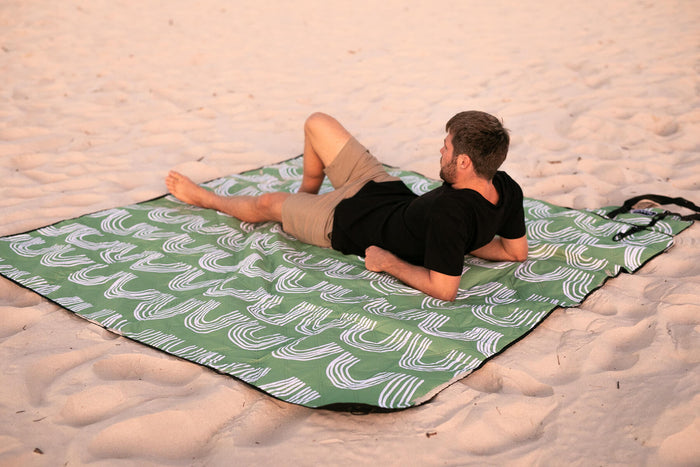 Green recycled picnic blanket with white geometric wave pattern, laid on beach sand with man resting on it.