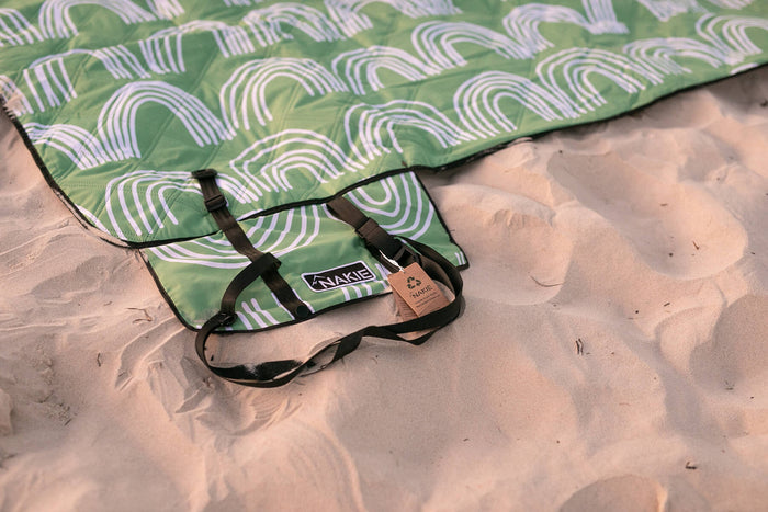 Green recycled picnic blanket with white and purple concentric arc pattern, black straps and carry handle on sand.