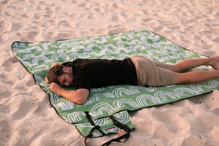 Green and white geometric patterned picnic blanket with black binding, laid flat on sand with person lying on it