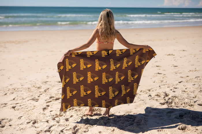 Brown beach towel with gold Hawthorn Hawks AFL logo pattern, held by person on sandy beach