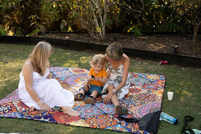 Colorful abstract patterned picnic blanket in use outdoors on grass with two adults and a child sitting on it.