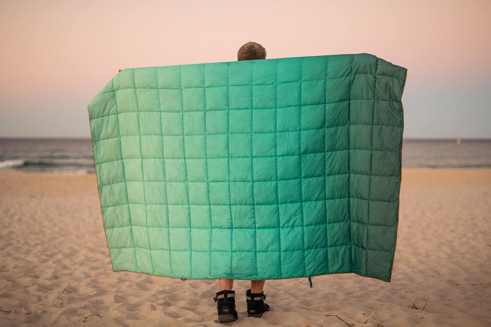 Green quilted puffy blanket held open on beach, showing box-stitch pattern and lightweight fill