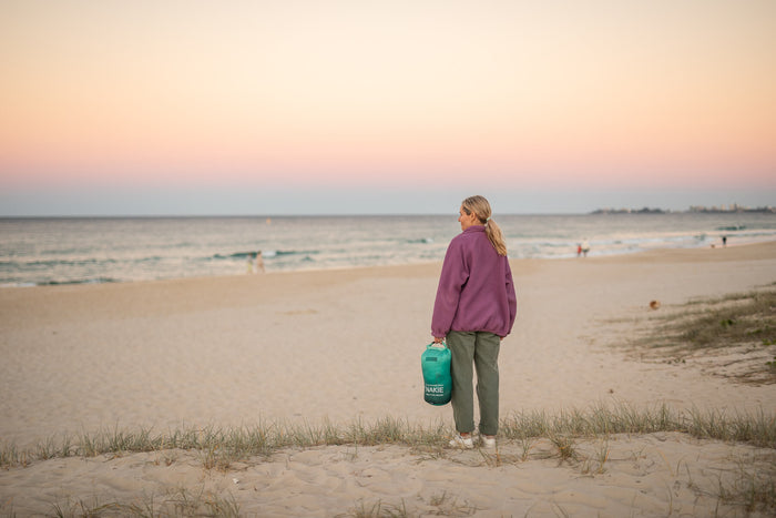 Woman holding green Nakie puffy blanket at beach at dusk, wearing purple jacket and olive pants
