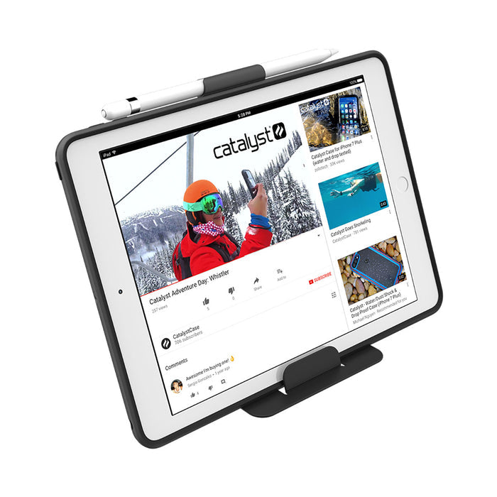 A white iPad (Gen 6/5), 9.7" in a Catalyst Case US Impact Protection Case stands upright with a stylus on top, displaying a YouTube video titled ÃƒÂ¢Ã¢â€šÂ¬Ã…â€œCatalyst Adventure Day: Whistler,ÃƒÂ¢Ã¢â€šÂ¬Ã‚Â featuring ski lift scenes and drop protection.