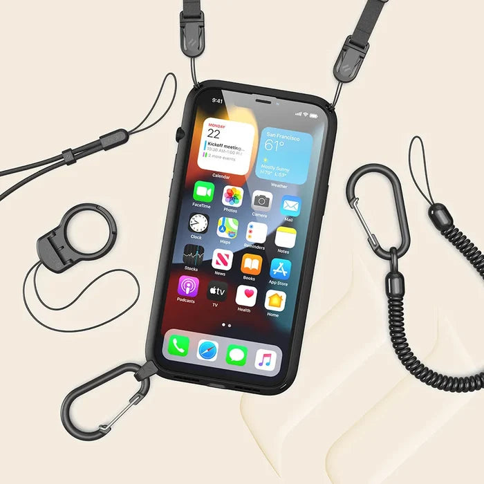 An iPhone 13 mini in a Catalyst Case US Influence Case is attached to a lanyard with adjustable clips and carabiners. The screen shows apps and weather, and accessories like a coiled strap and ring holder rest on a beige surface.