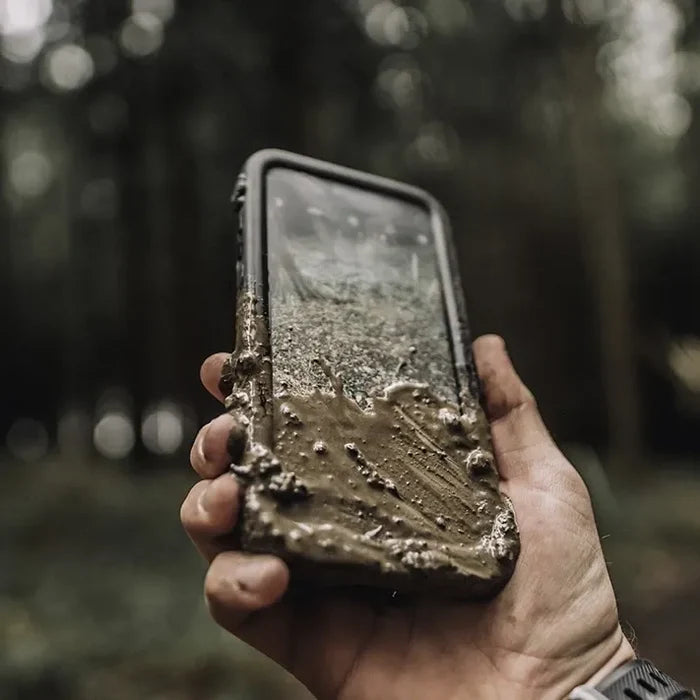 A hand holds an iPhone 14 Plus in a Catalyst Case US Waterproof Case, covered in wet mud on the lower half. The mostly clean screen shows some dirt. In the blurry forest background, the wearer sports a watch.