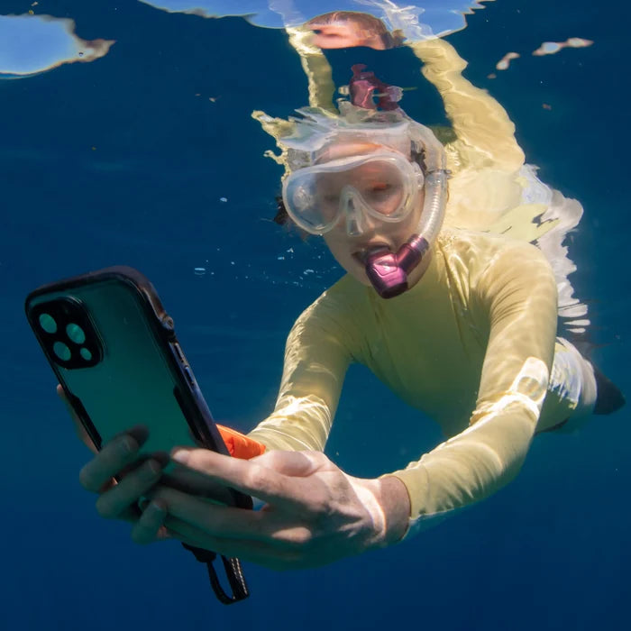 A swimmer in a yellow swimsuit, snorkel, and goggles glides underwater holding an iPhone 14 Plus protected by the Catalyst Case US Waterproof Case as sunlight streams through the blue water and their reflection shimmers above.