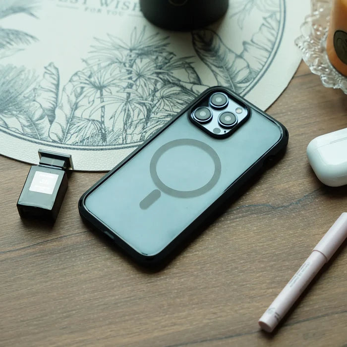 An iPhone 14 Pro Max in a clear Catalyst Case US Influence Case (MagSafe compatible) rests on a wooden table beside a black perfume bottle, white wireless earbud case, pink pen, and a round botanical placemat.