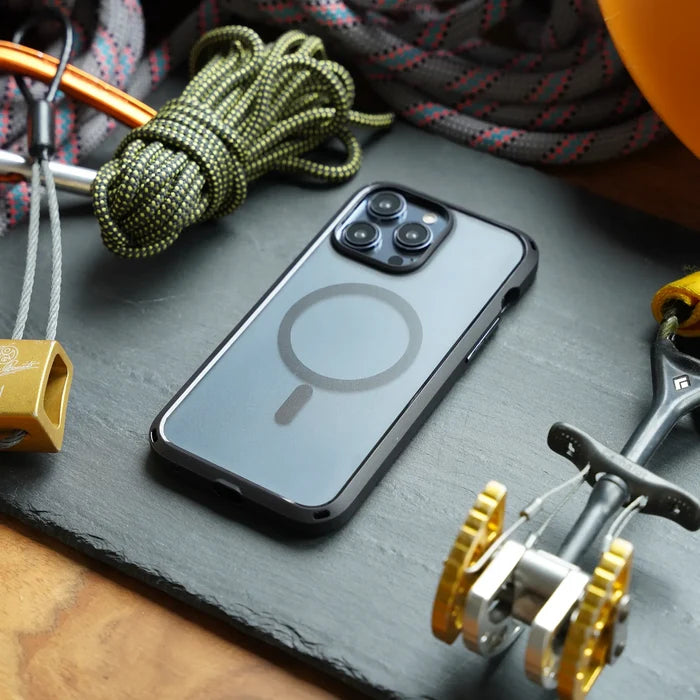An iPhone 15 Pro with the Catalyst Case US Influence Case (MagSafe compatible) rests on a dark slate surface, surrounded by climbing gear—carabiners, a coiled rope, a cam device, and part of a yellow helmet—with focus on its MagSafe circle.