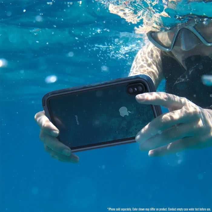 A person in swim goggles holds an iPhone Xs Max encased in a Catalyst Case US Waterproof Case underwater in a clear blue pool, sunlight streaming from above and bubbles surrounding their hands.