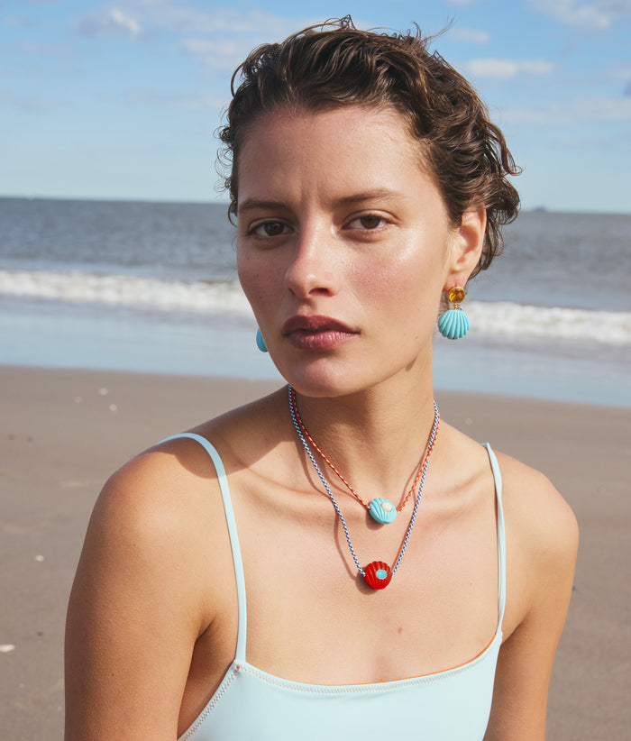 Model on beach wears blue swimsuit with Kai Necklaces in Ultramarine and Red Hot and Kai Earrings