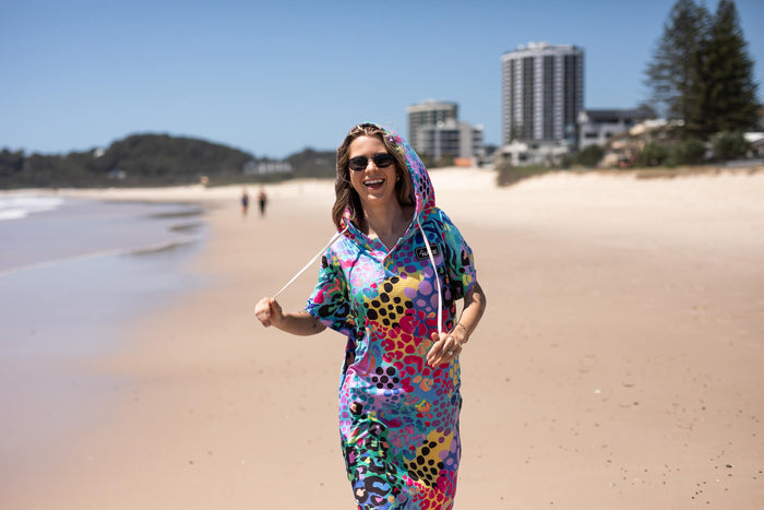 Hooded towel in rainbow multicolor geometric print with hood up, worn on beach by woman wearing sunglasses