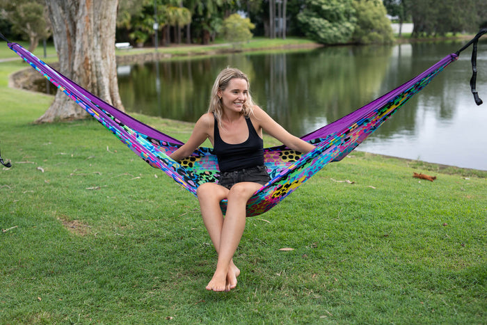 Recycled hammock with rainbow geometric pattern and purple edges, displayed outdoors between trees.