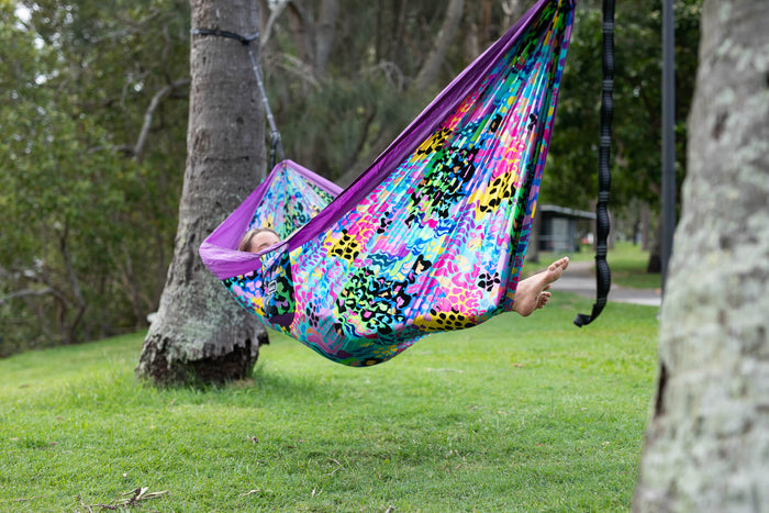Colorful recycled hammock with rainbow leopard print and purple trim, suspended between trees with black straps.