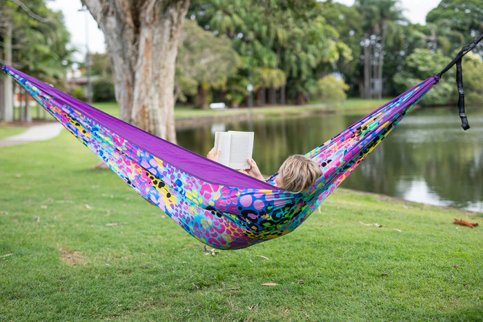 Purple and multicolored patterned recycled hammock with black straps, suspended between trees by a waterside park.