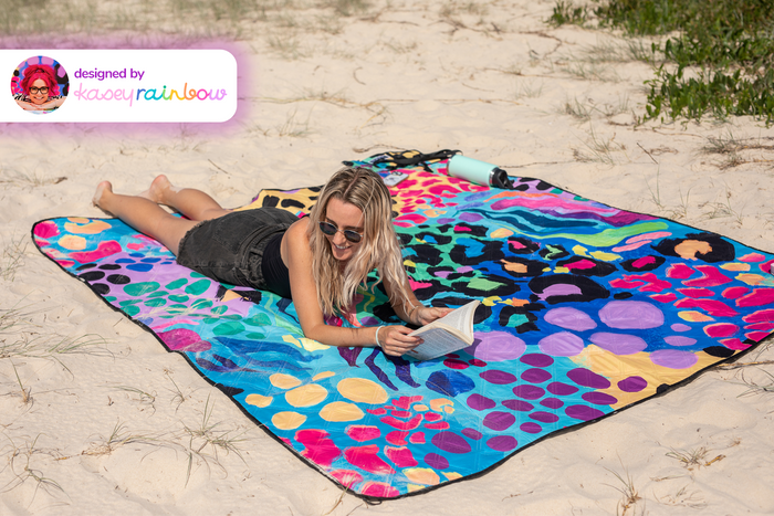 Colorful recycled picnic blanket with multicolored leopard and geometric patterns, laid on sand with woman reading.