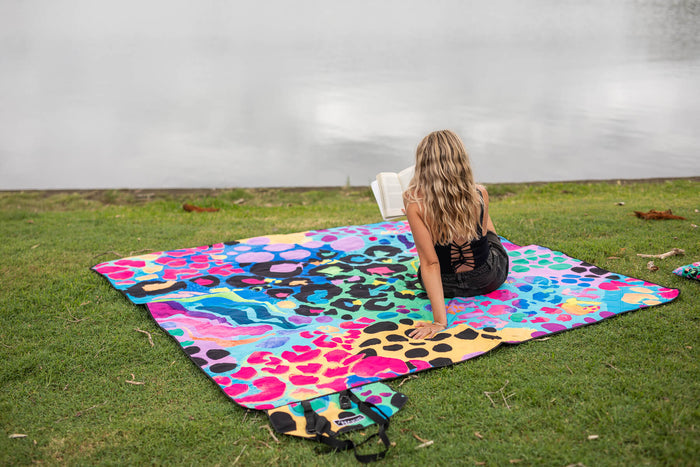 Colorful recycled picnic blanket with multicolor animal print pattern spread on grass, woman reading on blanket.