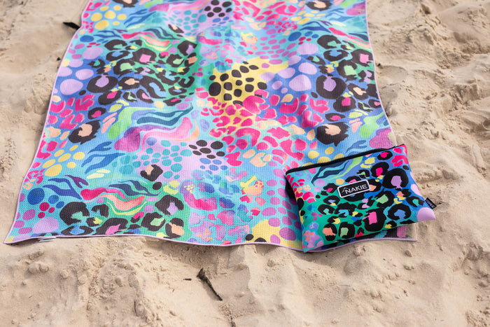 Multicoloured recycled sand-free beach towel with abstract leopard and wave pattern, displayed on sand with matching pouch.