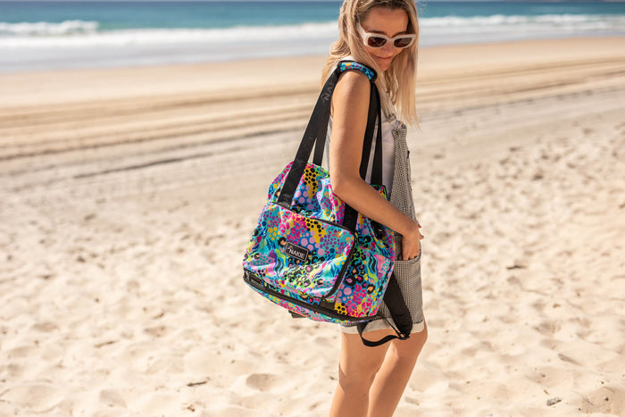 Colorful recycled tote bag with rainbow abstract pattern and black straps, worn on shoulder at beach.