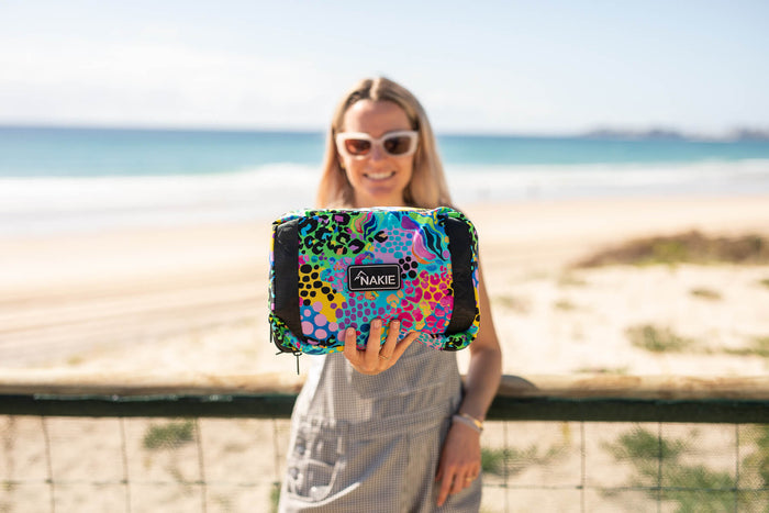 Colorful abstract patterned recycled tote bag with black panels and Nakie label, held at beach