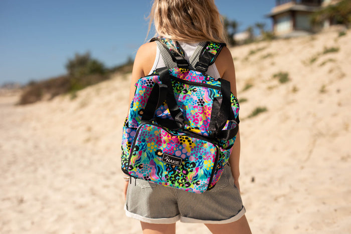 Multicolored neon geometric patterned tote backpack with black straps worn on shoulders at beach.