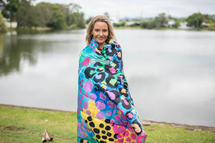 Sustainable down puffy blanket in multicolour leopard print wrapped around person by water
