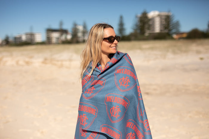 Blue beach towel with red Melbourne Demons AFL logo pattern, draped around woman on sandy beach
