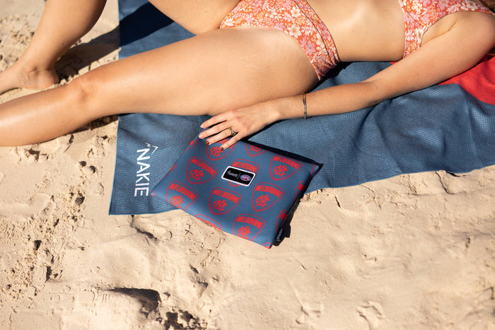 Melbourne Demons navy and red patterned recycled beach towel laid on sand with people nearby.