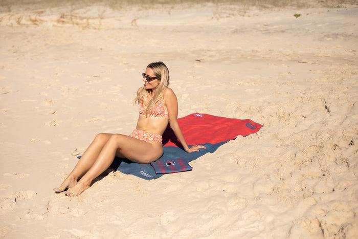 Red and navy AFL beach towel with Melbourne Demons branding, displayed on sand at beach