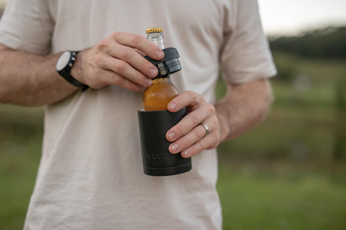 Black neoprene bottle cooler holding amber beer bottle with gold cap, worn on outdoor setting