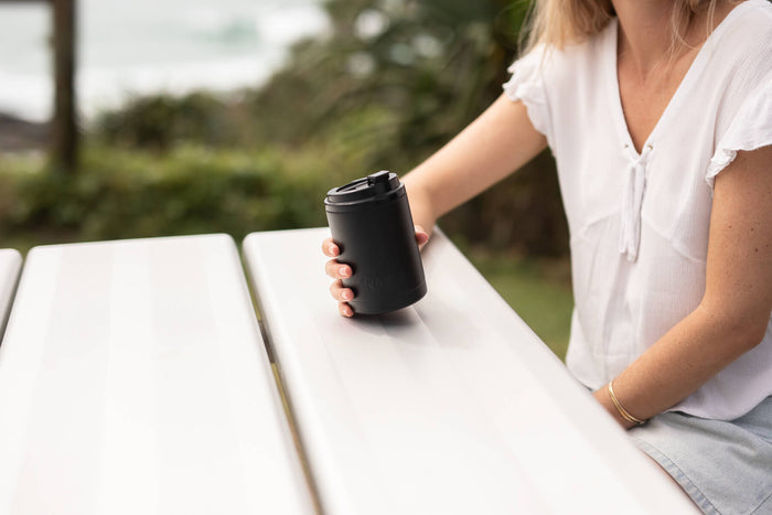 Black insulated coffee cup lid held in hand, outdoor garden setting
