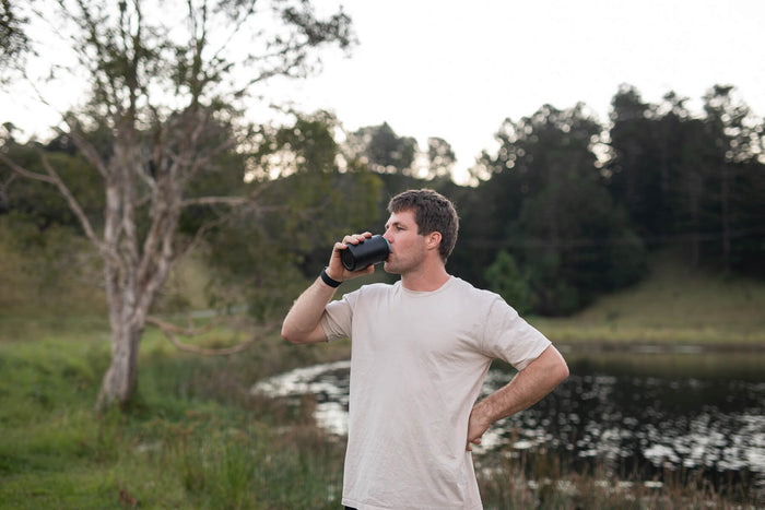 Midnight black insulated coffee cup lid shown in use outdoors by a man drinking from a black beverage container near a river