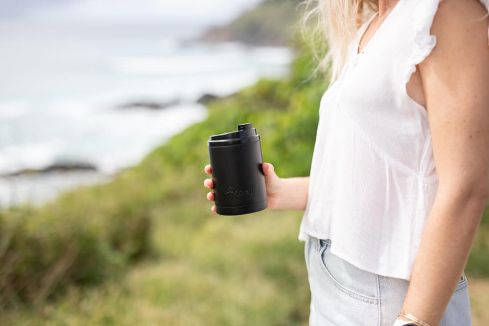Black insulated coffee cup with lid held in hand, outdoor coastal setting