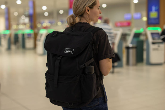 Black recycled travel backpack with flap closure and side pockets, worn on model's back in airport setting