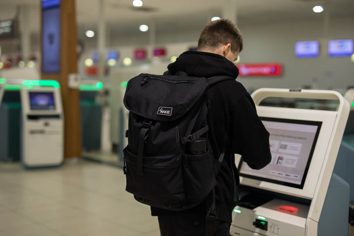 Black recycled travel backpack with front flap pocket and side straps, worn by person in airport setting