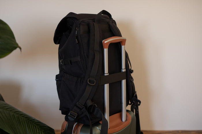 Black recycled travel backpack with tan leather accents, shown attached to luggage handle with multiple zippered pockets.