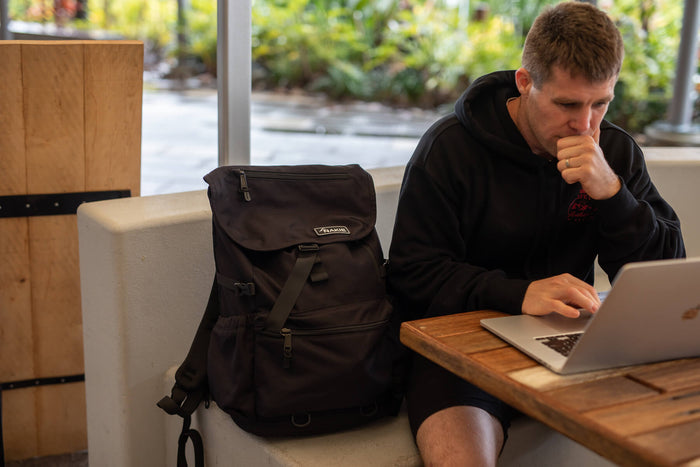 Black recycled travel backpack with top and side zip pockets, padded straps, displayed on seat next to laptop.