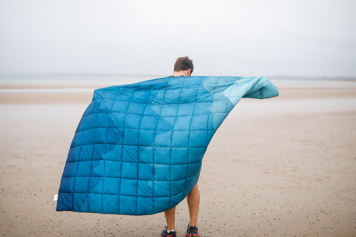 Blue quilted puffy blanket worn over shoulders on beach, showing lightweight insulated design