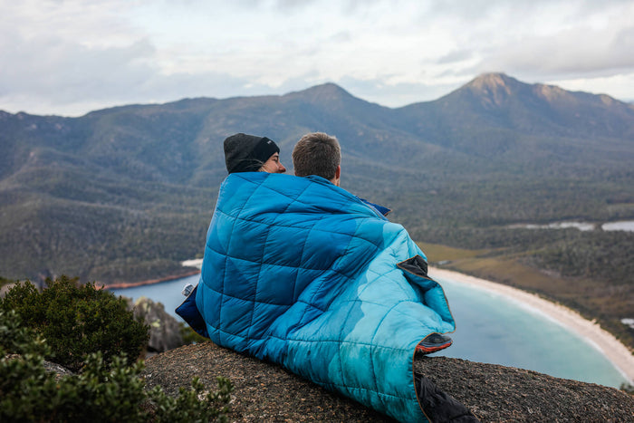 Blue quilted down puffy blanket wrapped around two people sitting on mountain overlook